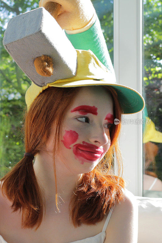 14 / 15岁的红发少女坐在阳光明媚的温室里玩气球爆破游戏，玩气球爆破游戏，戴着小丑脸和傻帽